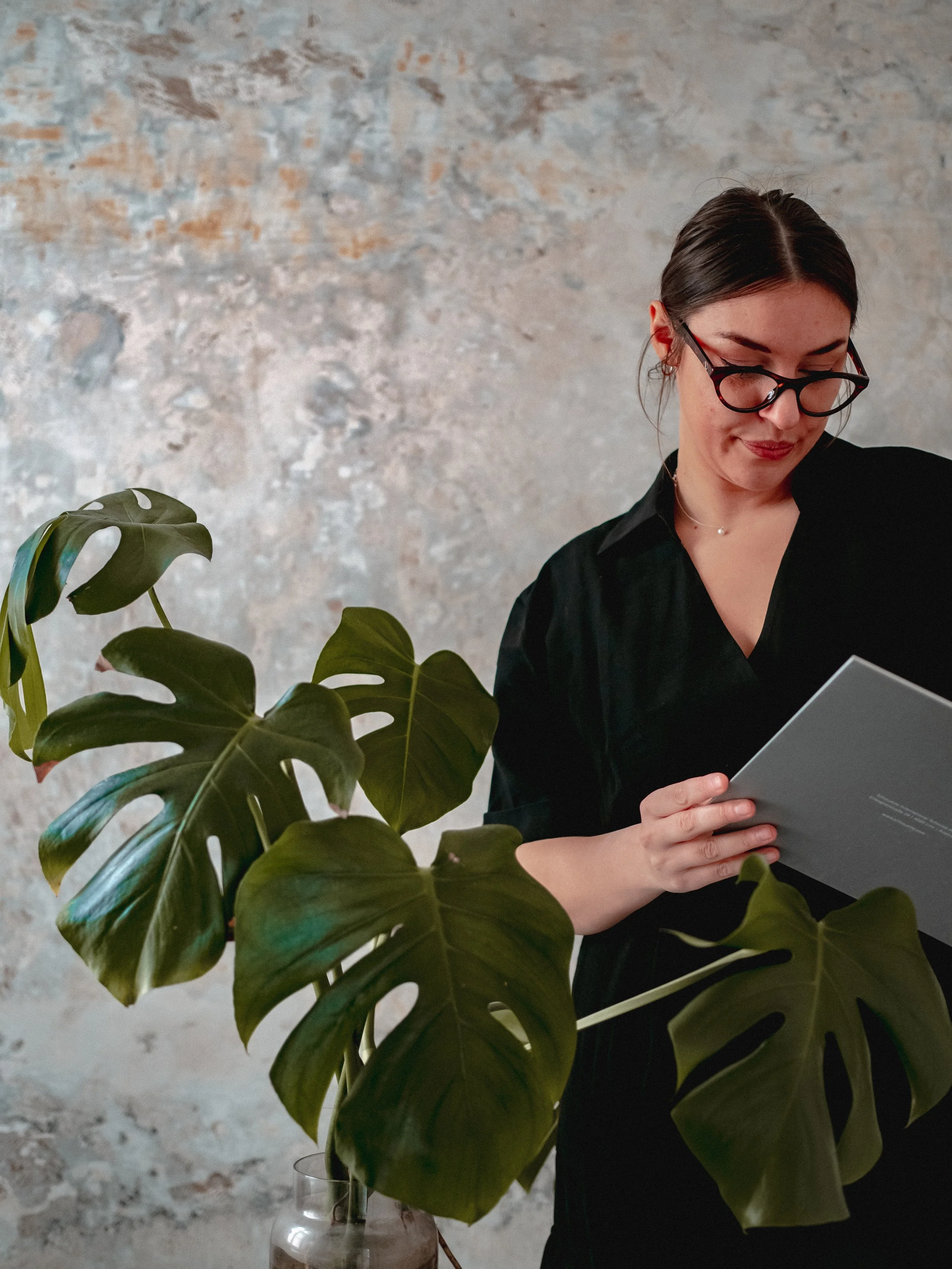 A woman with dark hair and glasses reads a book or magazine next to a large monstera plant against a textured wall background.