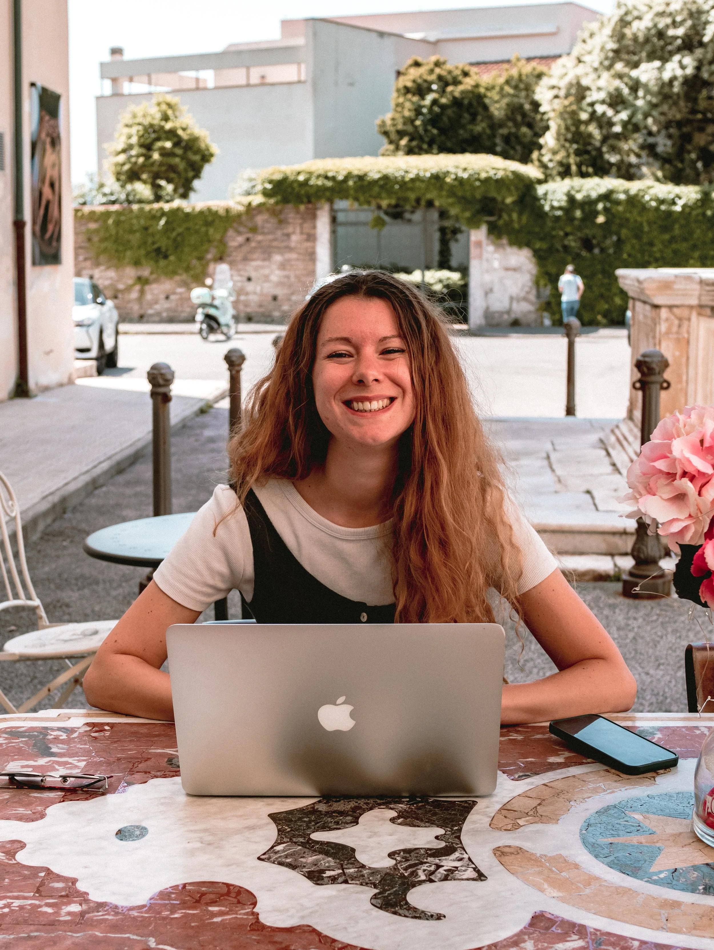 A young woman with long, curly, reddish-brown hair smiling at a table outdoors with a MacBook, smartphone, and pink flowers, in a sunny urban setting with trees and buildings in the background.