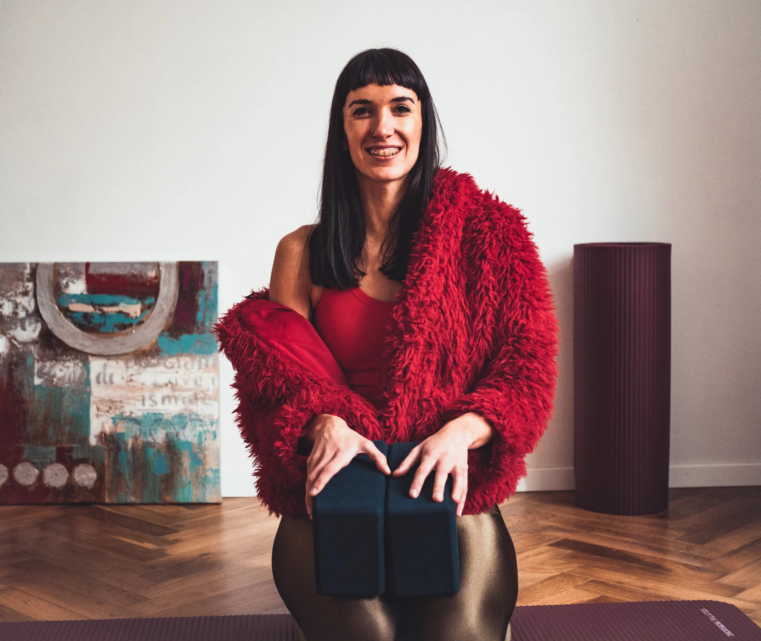 Woman in a red fuzzy jacket and red top holding yoga blocks on a yoga mat in a room with wooden flooring, a painting on the wall, and a tall red speaker.