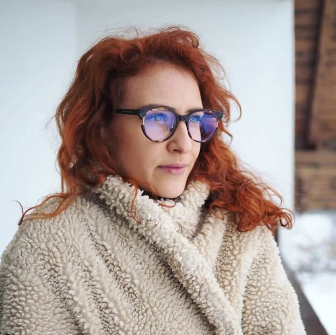 Woman with red curly hair and glasses wearing a beige fleece coat outdoors in winter.