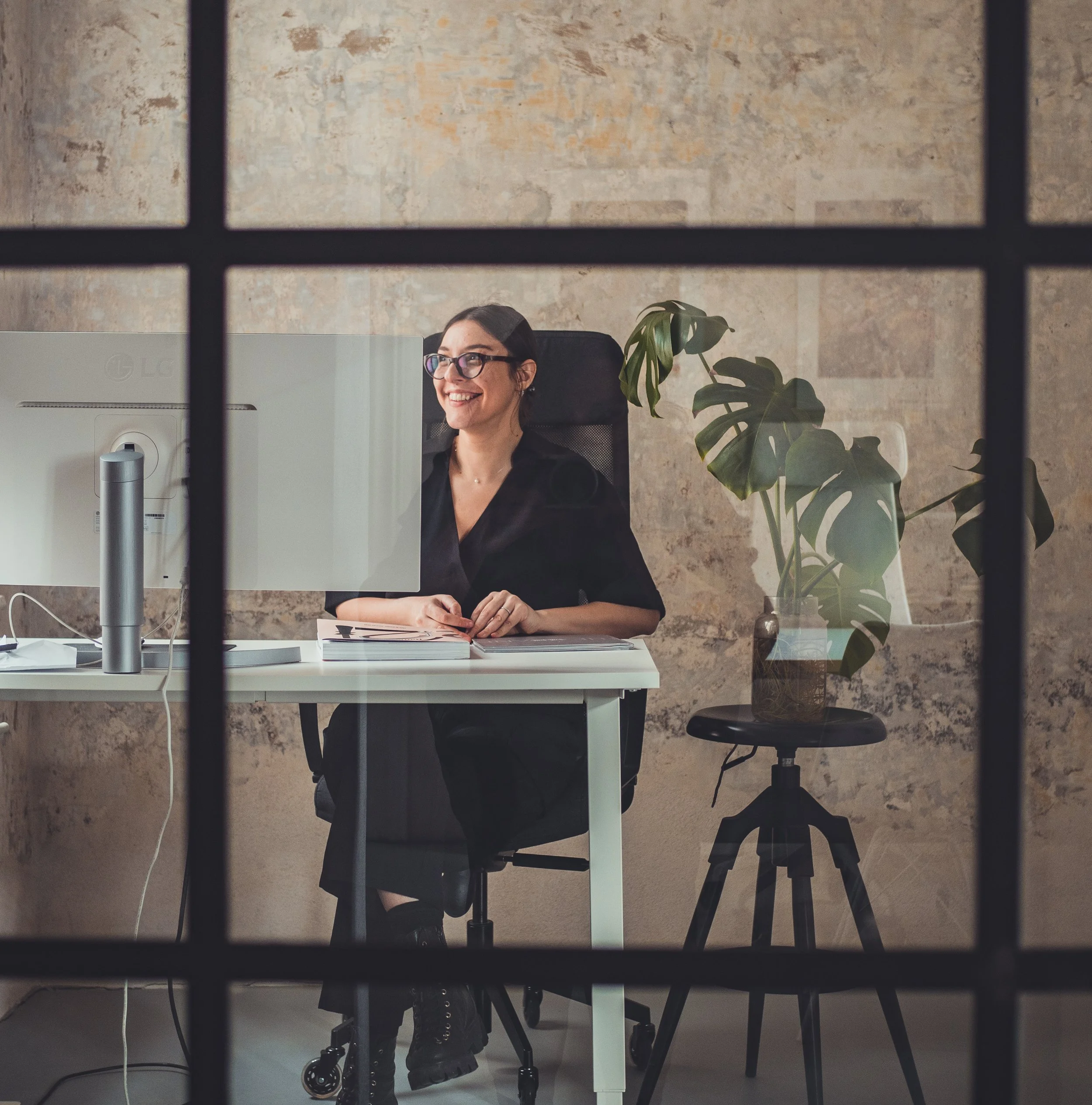 A woman with glasses sitting at a desk and smiling, viewed through a glass window with black grid pattern, with a computer, notes, and green plants in the background.