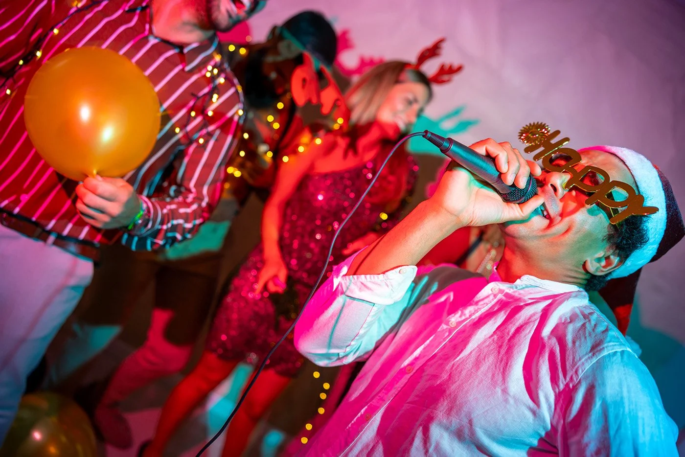 Four adults enthusiastically singing karaoke at a Christmas party. The singer in the foreground wears a Santa hat and novelty glasses, holding a microphone.