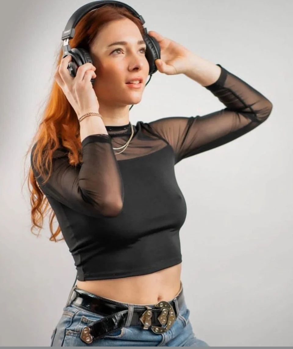 A young woman with red hair wearing black sheer long-sleeve top and blue jeans, listening to music with large over-ear headphones, standing against a plain background.