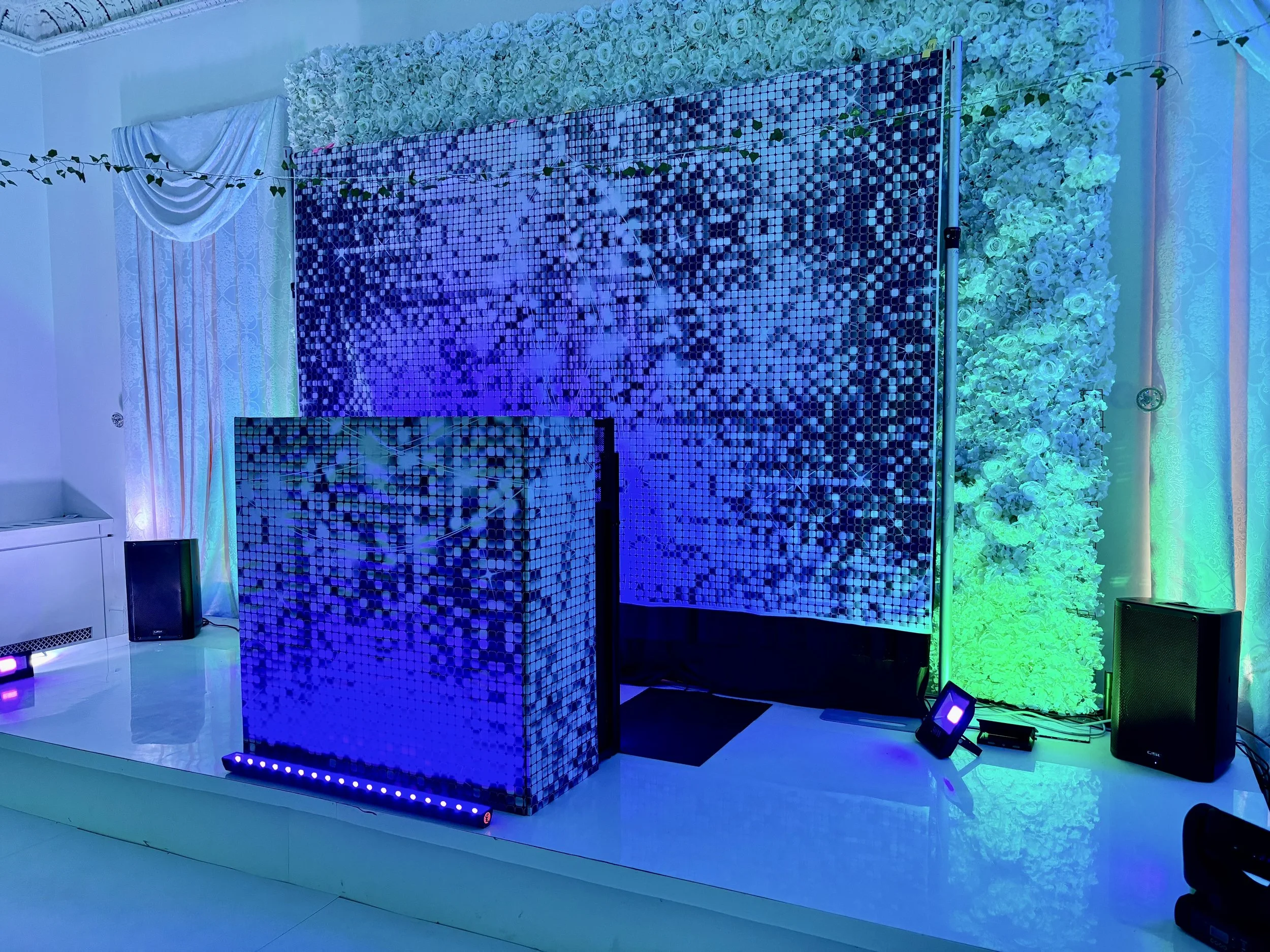 Decorative event backdrop with white flowers, green lighting, and a mosaic mirror DJ booth, with speakers and LED lights arranged in a celebration or party setting.