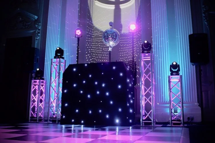 DJ booth with black tablecloth illuminated with blue and purple LED lights, four purple-lit truss towers with moving head lights, a disco ball hanging from high ceiling, and a checkered dance floor in an elegant room with tall white columns.