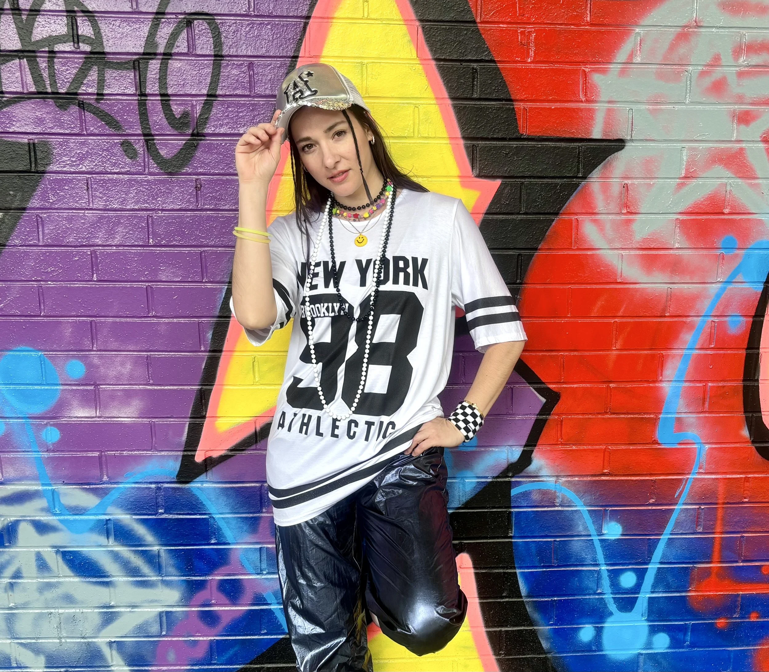 Four young women and one young man singing and dancing with a microphone in a bright room with white brick walls.