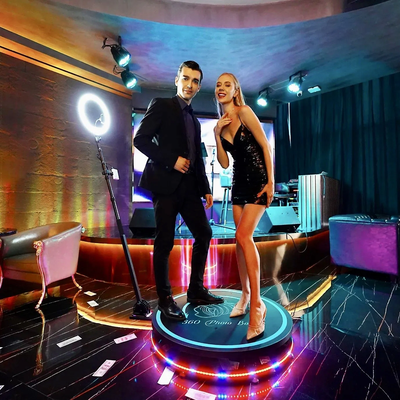 A man in a black suit and a woman in a sparkly black dress standing on a 360-degree photo booth platform in a dimly lit nightclub or event space.