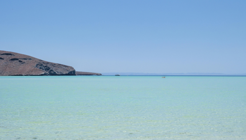 A calm, turquoise sea with a distant boat and a arid, rocky coastline under a clear blue sky.