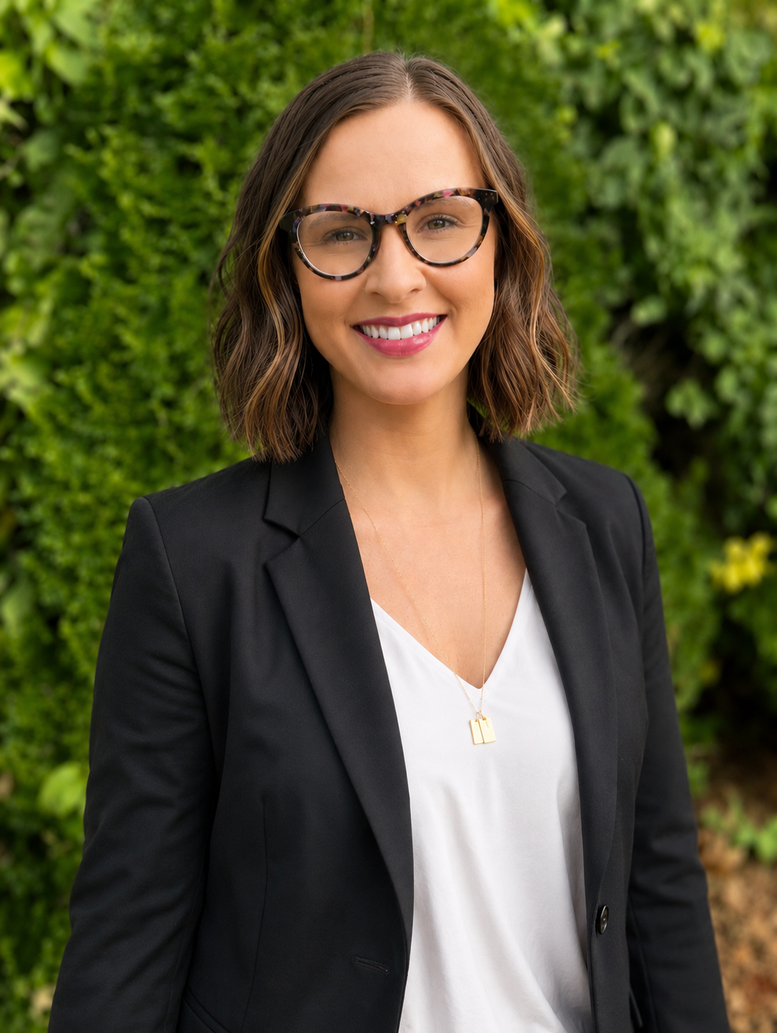 Woman with long brown hair, wearing glasses, a black jacket, and a gray top, standing outdoors with green foliage in the background.