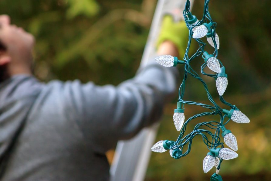 Person taking down outdoor Christmas lights from a ladder during daylight. Removal of seasonal holiday lighting.