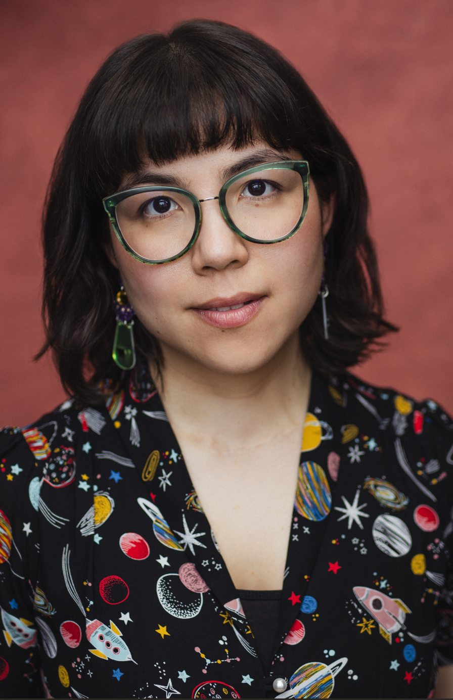 Emi's headshot, a mixed East Asian woman in her early 30s with a bob and a fringe, wearing large glasses, space-themed earrings and dress, looking at the camera with a slight smile