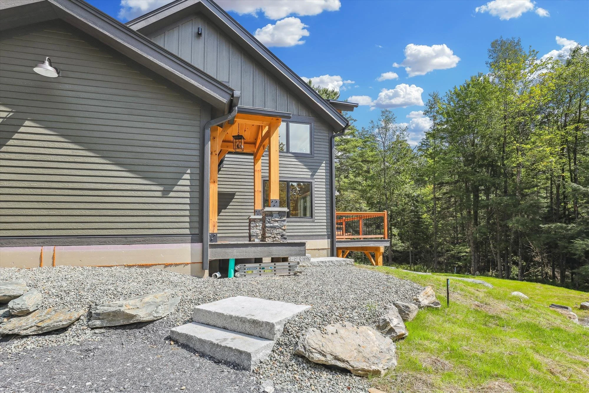 Side view of a house under construction with new steps, gray siding, wood accents, and a deck, surrounded by trees on a sunny day.