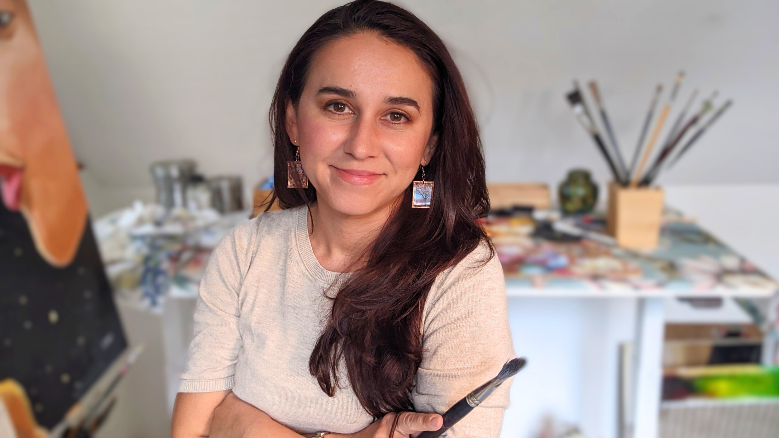 A picture of the artist and oil painter Adrienn Baris in her studio, holding a paintbrush and smiling, with art supplies and paintings in the background.