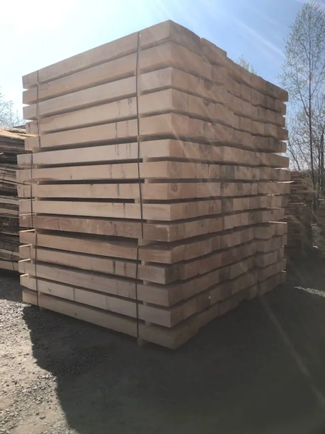 Large stack of wooden planks outdoors on a gravel surface, with trees and a blue sky in the background.
