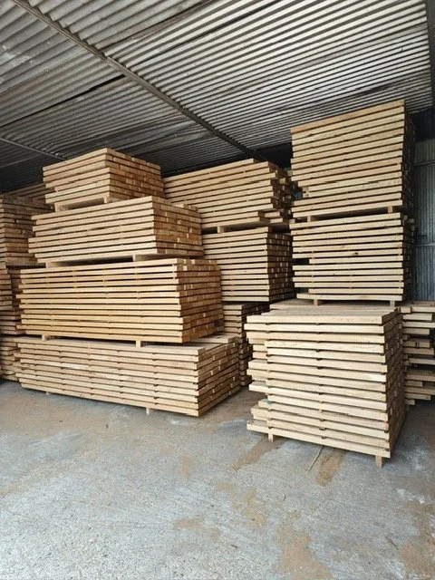 Stacks of wooden planks stored in a warehouse with a corrugated metal roof.