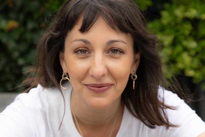 A woman with short brown hair and gold hoop earrings smiling at the camera outdoors.