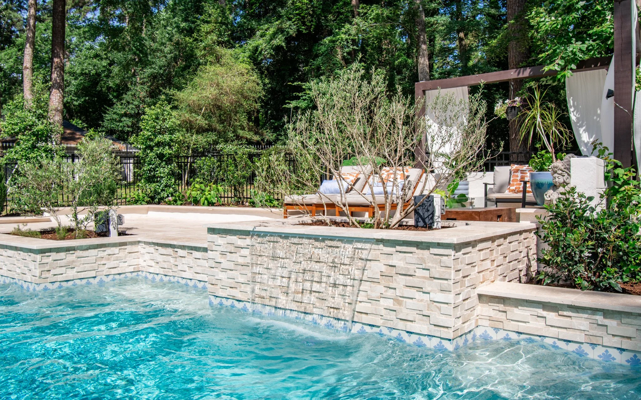A backyard with a swimming pool, stone patio, and outdoor seating area with cushions and decorative pillows. There are trees and greenery in the background, and a fountain feature on the wall of the patio.