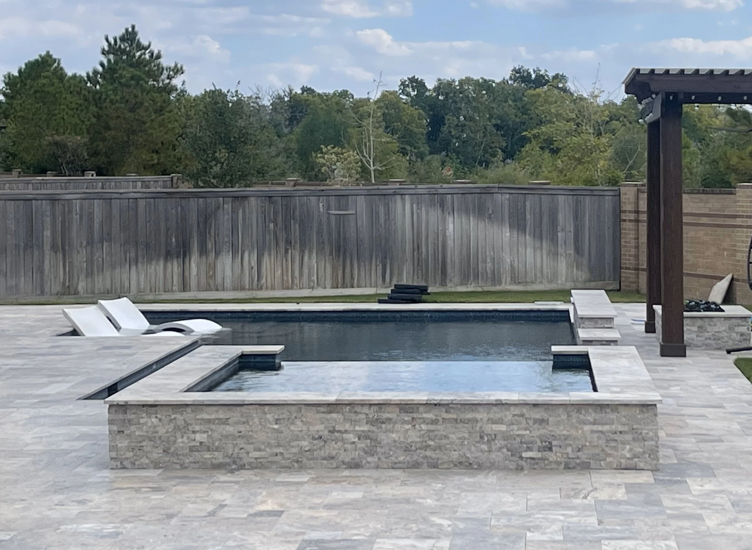 A backyard scene with a swimming pool, two white lounge chairs, a wooden fence, and green trees in the background under a partly cloudy sky.