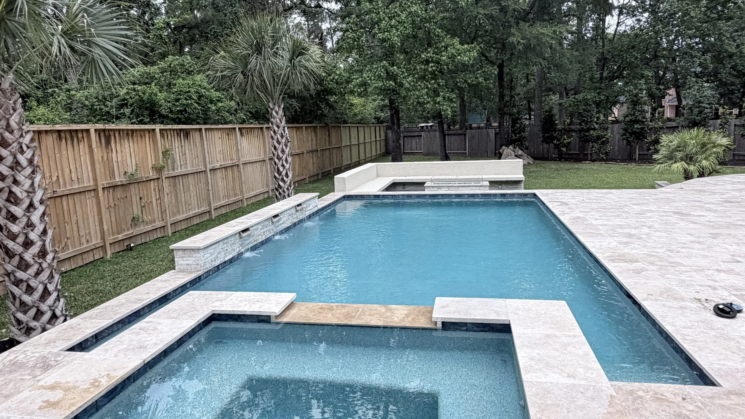A backyard swimming pool with bordered by stone tiles, surrounded by a wooden privacy fence and lush greenery including palm trees and other plants.