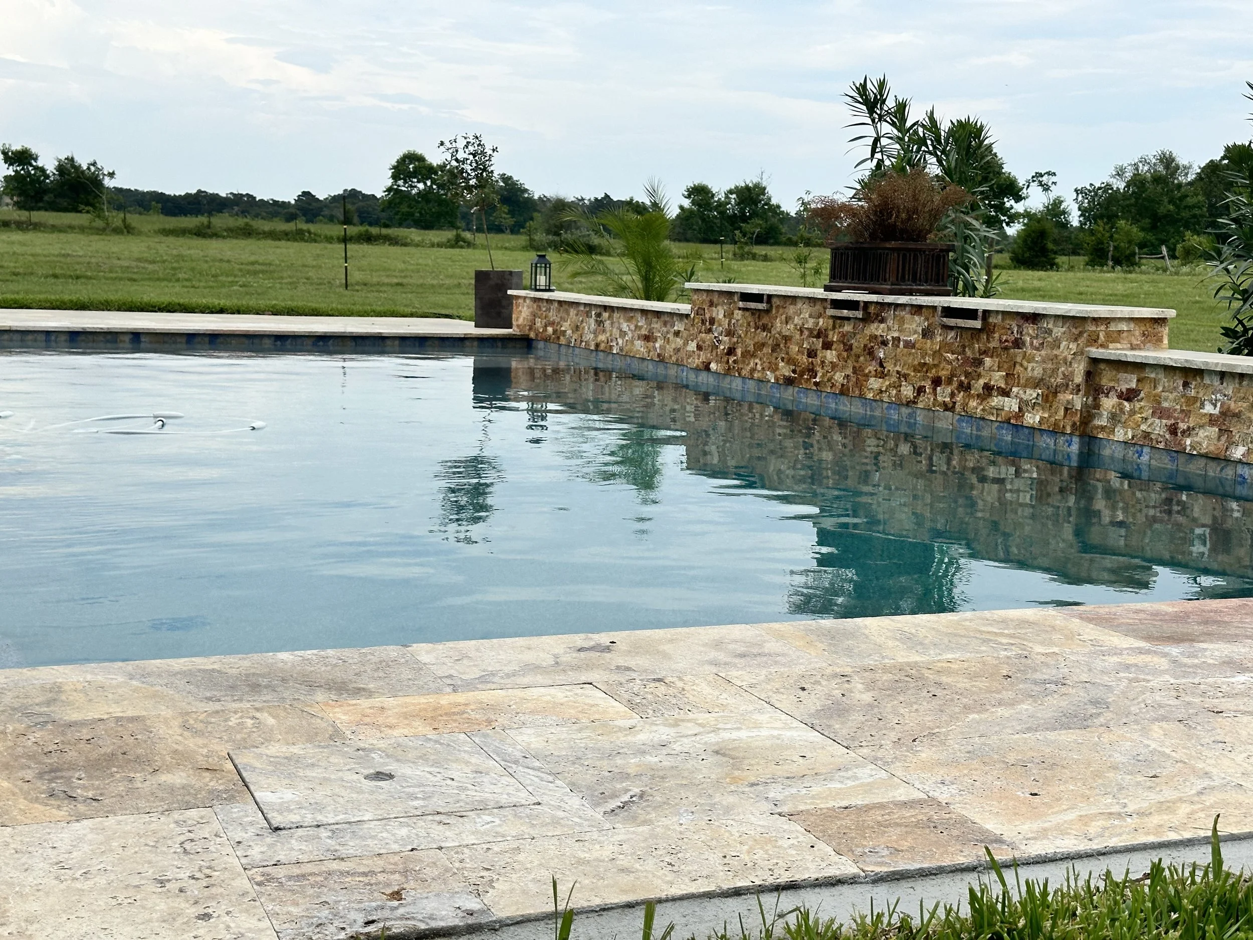 A swimming pool with a stone tile deck and a stone wall at the edge, situated in a grassy field with trees in the background, under a cloudy sky.