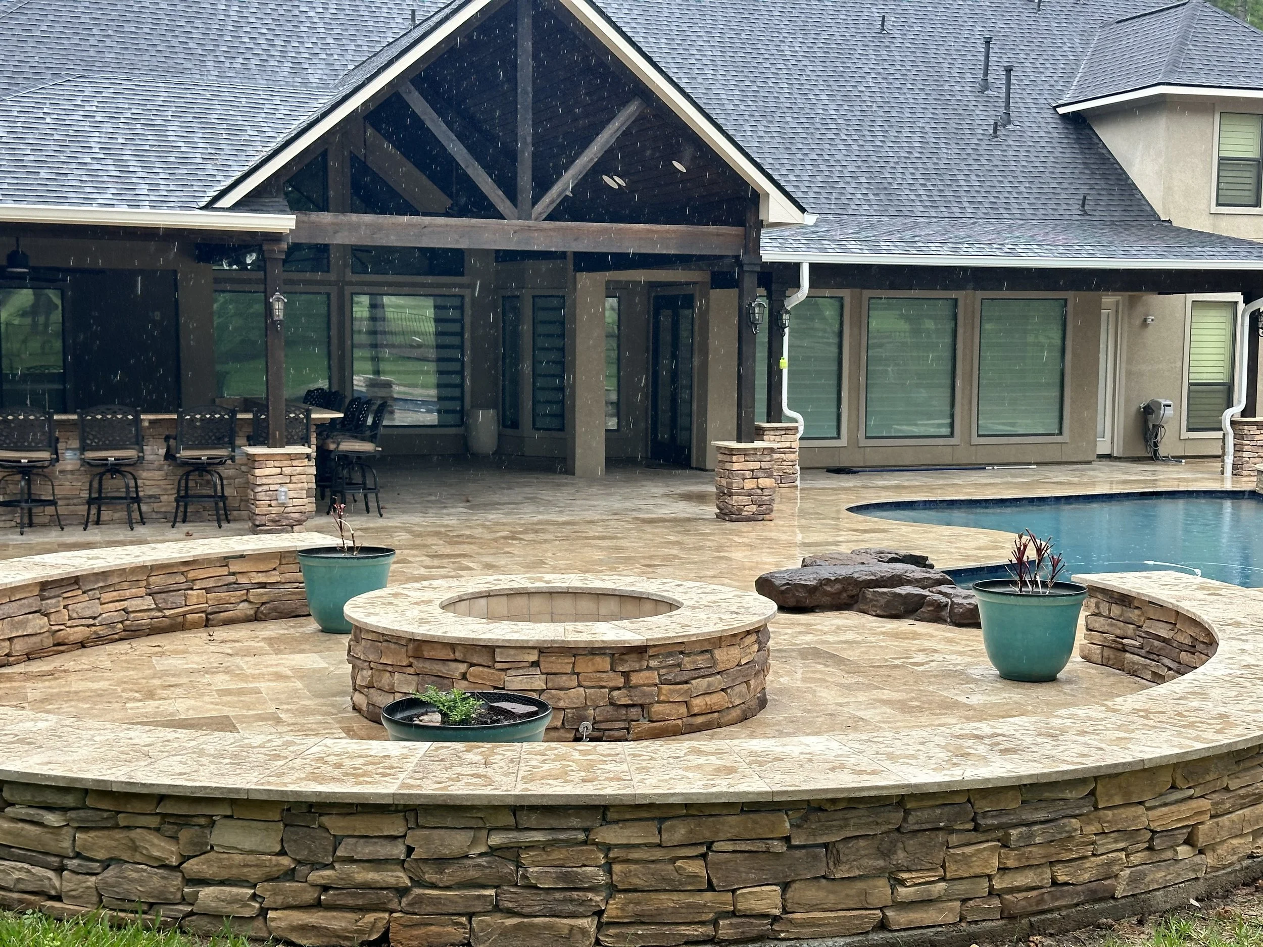 A backyard patio with a stone fire pit, potted plants, outdoor seating, a swimming pool, and a house with a covered porch and large windows. Rain is falling.
