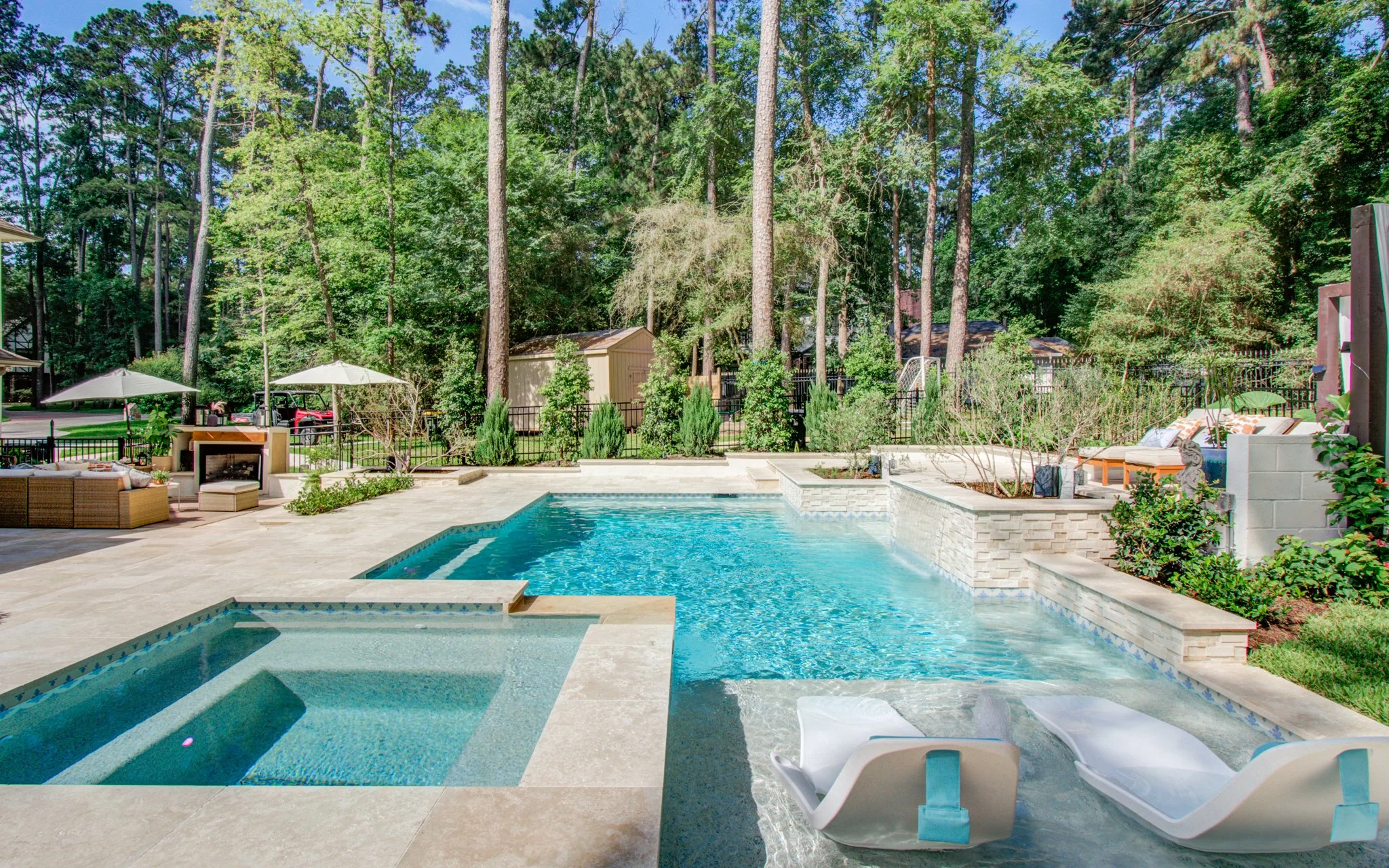 A backyard swimming pool with clear blue water, surrounded by a concrete deck and white lounge chairs. In the background, there's a lush green forest of tall trees, some with umbrellas and outdoor furniture, creating a relaxing outdoor setting.