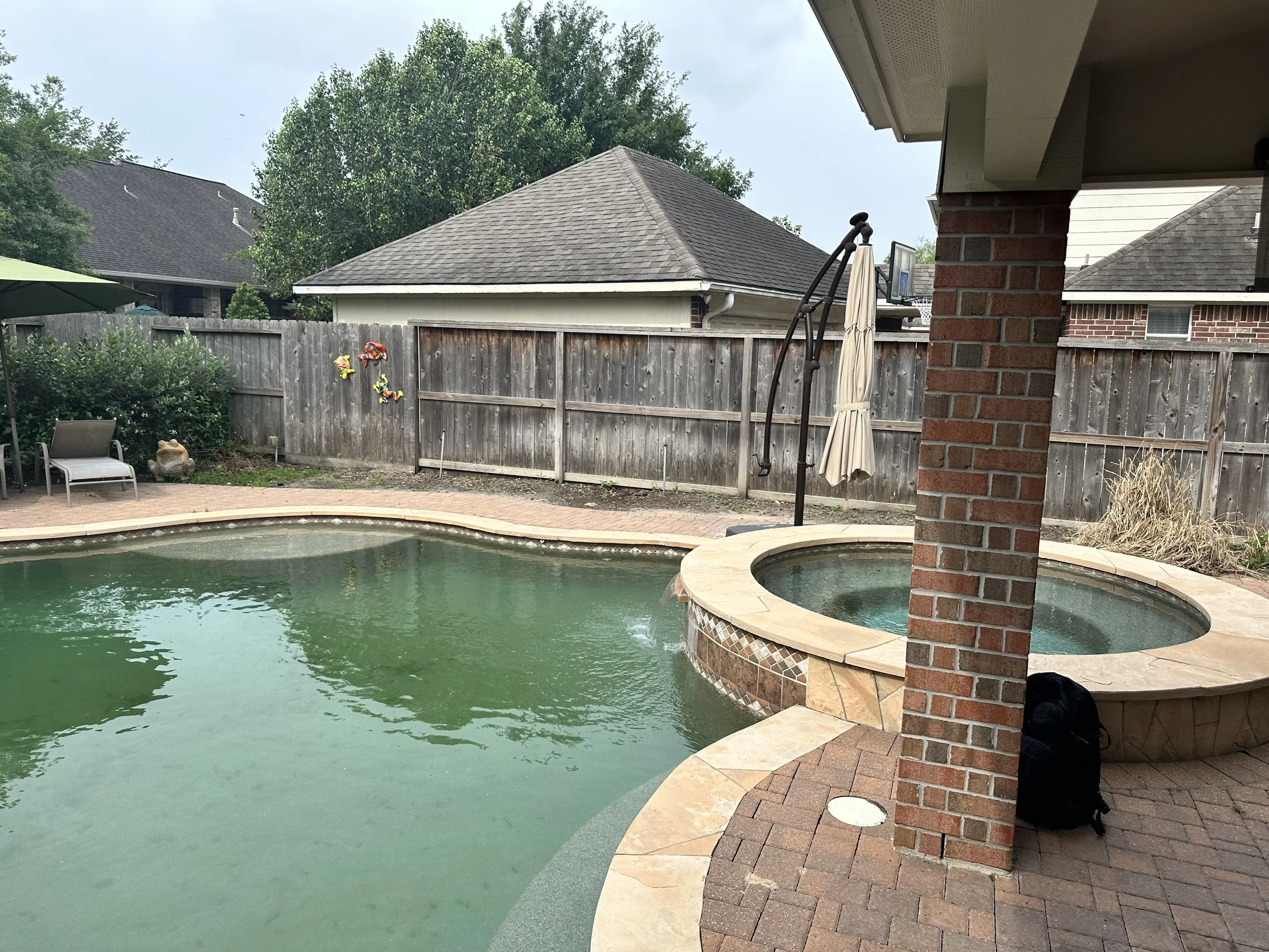 Backyard swimming pool with patio furniture, a umbrella, a wooden fence, a shed, and trees in the background.