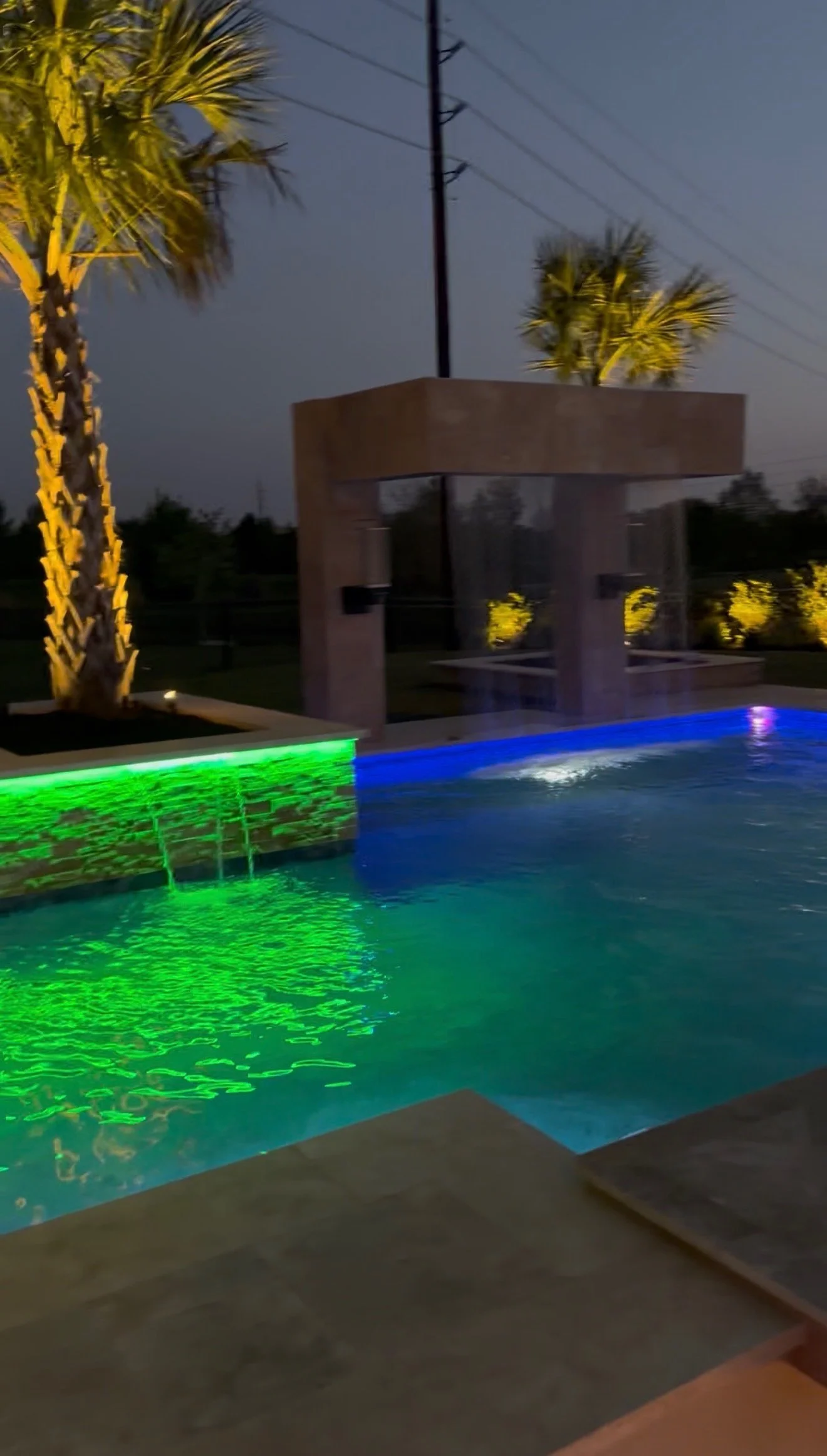 Night view of a swimming pool with green and blue underwater lights, surrounded by palm trees and modern architectural structures.