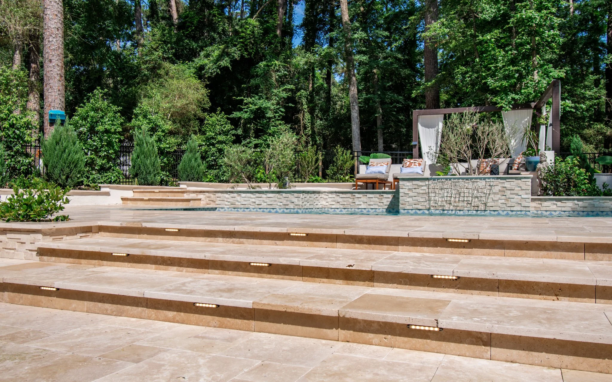 Outdoor patio with multi-level beige stone steps, a swimming pool at the bottom, and a seating area with outdoor furniture and cushions, surrounded by lush green trees.