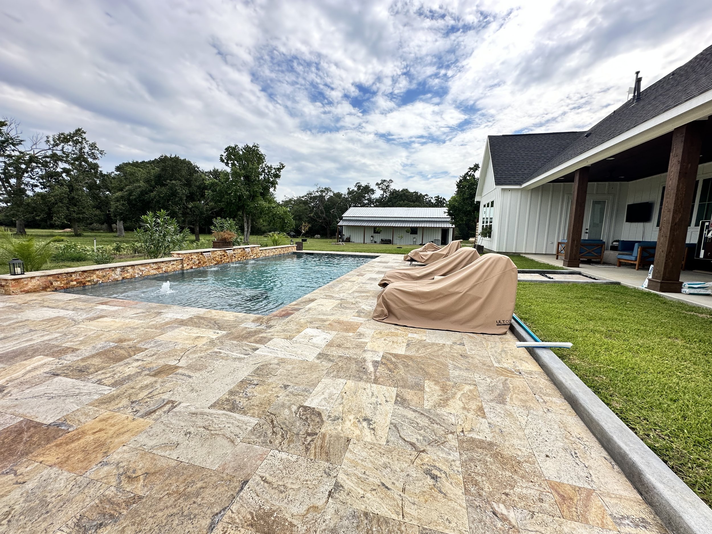A backyard scene featuring a swimming pool with a stone edge, beige lounge chairs covered with beige covers, a white house with a dark gray shingle roof, a large covered porch, green grass, a white shed, and trees under a partly cloudy sky.