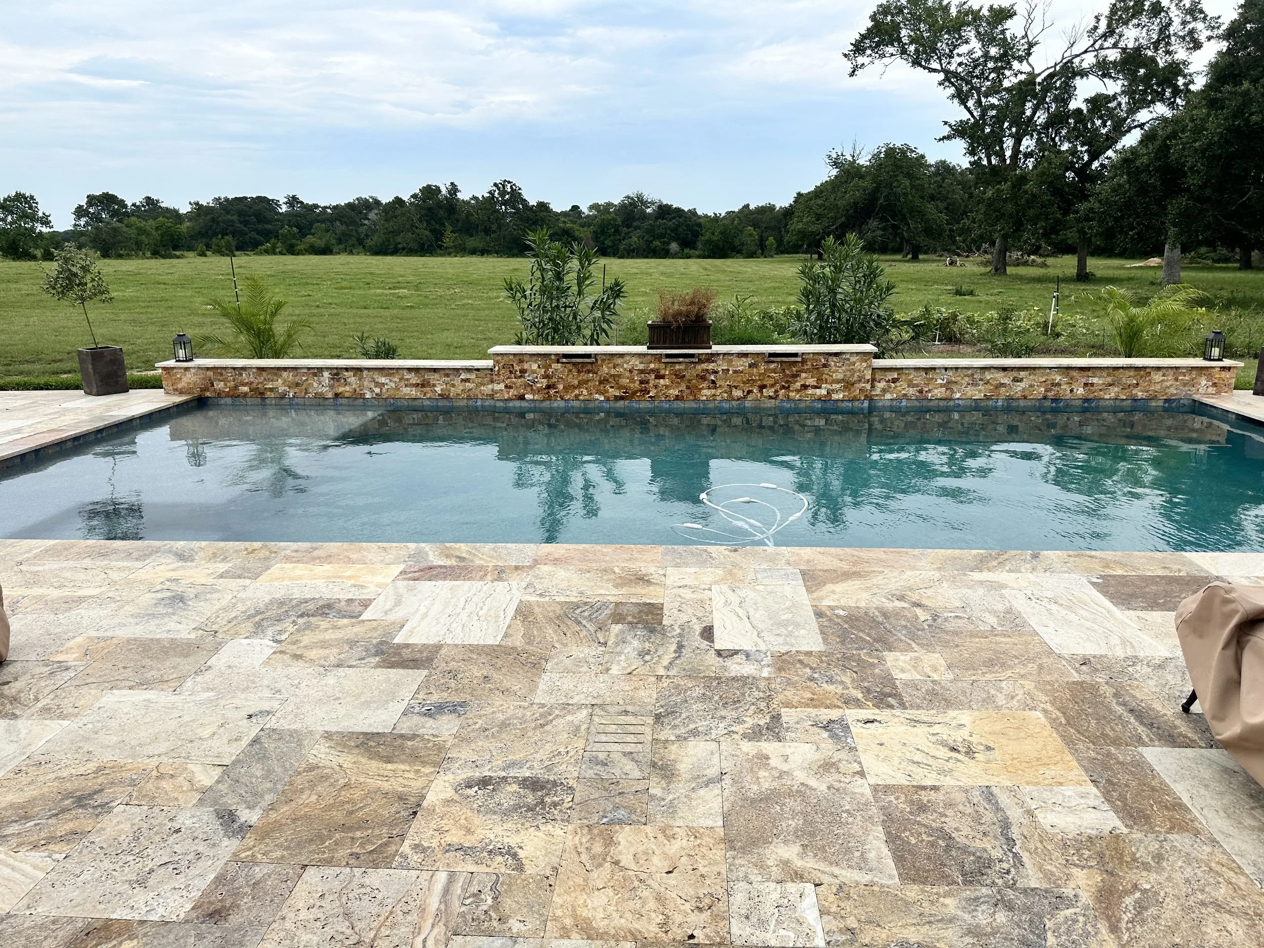 A backyard patio with a swimming pool, stone tile flooring, potted plants, and a grassy field with trees in the background under a cloudy sky.