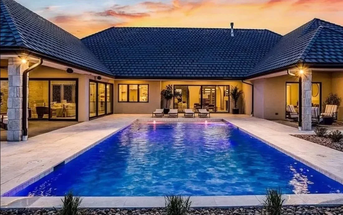 A backyard scene showing a modern house with a swimming pool, lounge chairs, and patio furniture during sunset