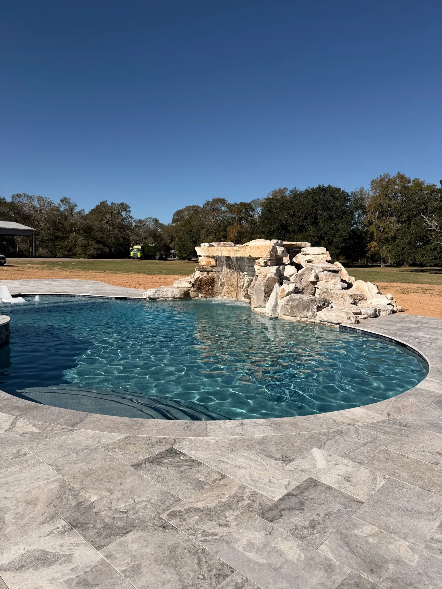 A backyard swimming pool with a water feature made of rocks and a waterfall, surrounded by a gray tile deck, with trees and a clear blue sky in the background.