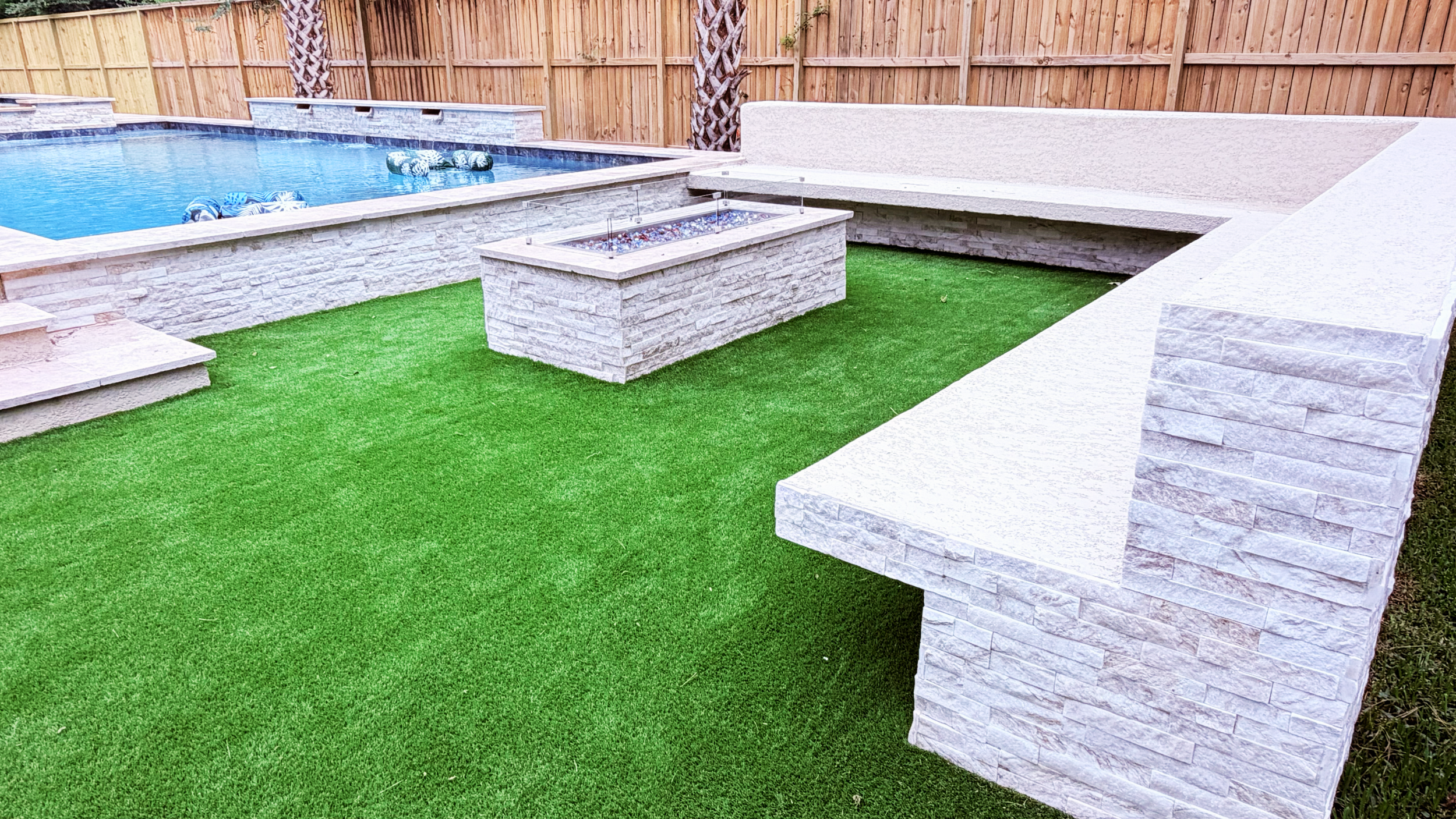Stone bench seating and fire pit on turf beside a pool in The Woodlands, TX