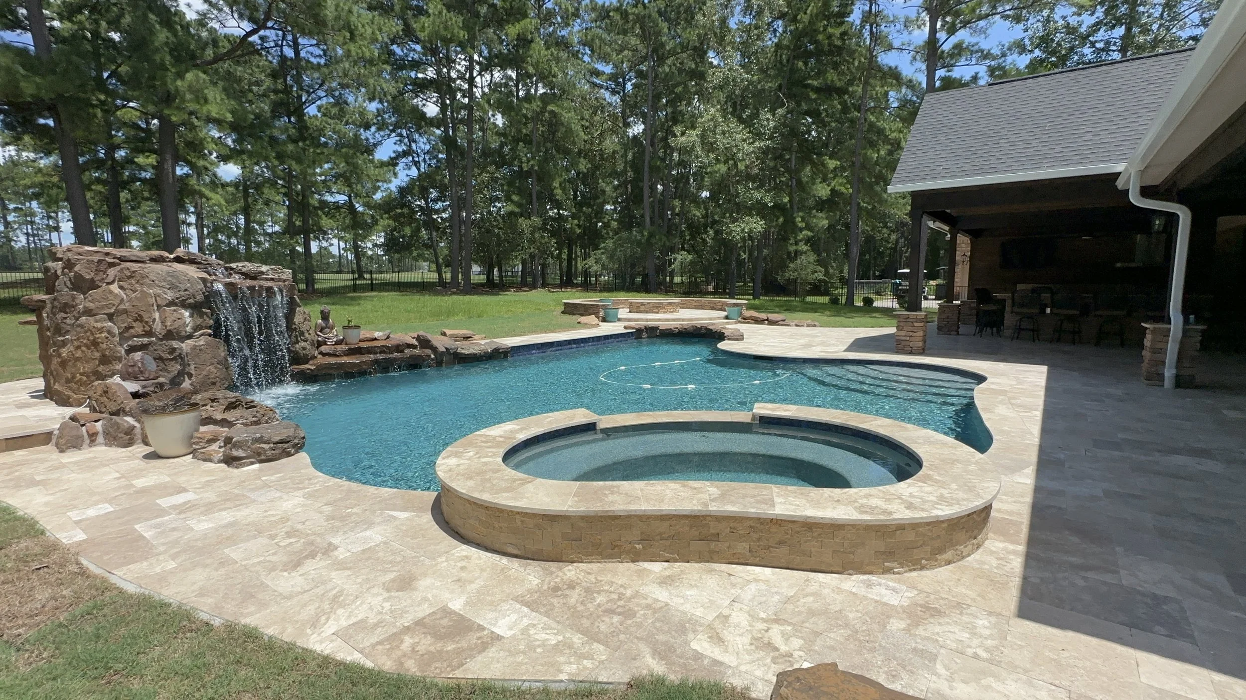 Backyard with swimming pool and hot tub, fountain feature with water cascading over rocks, surrounded by a stone patio, trees and a house with a covered patio area and outdoor furniture.