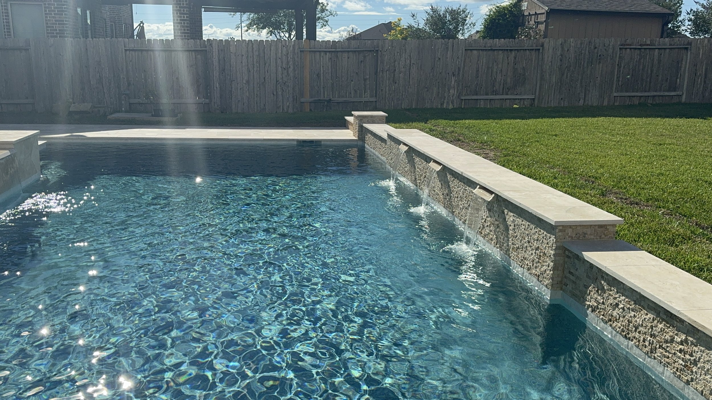 Backyard pool with water features, surrounded by a stone deck and a grassy yard, enclosed by a wooden fence. Sunlight reflects off the water's surface, with trees and houses visible beyond the fence.