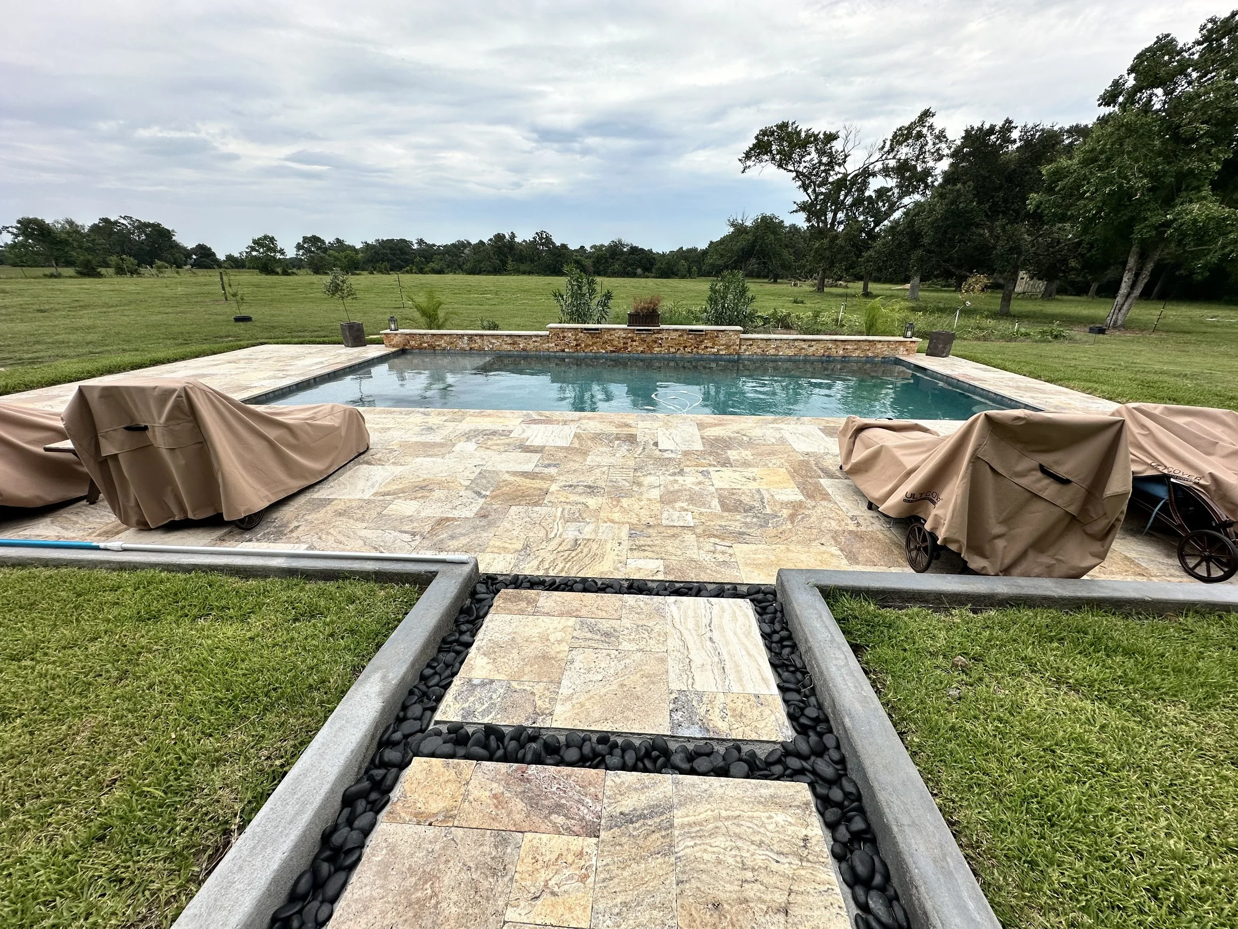 View of a backyard with a swimming pool, stone patio, and a lush green field with trees in the background.