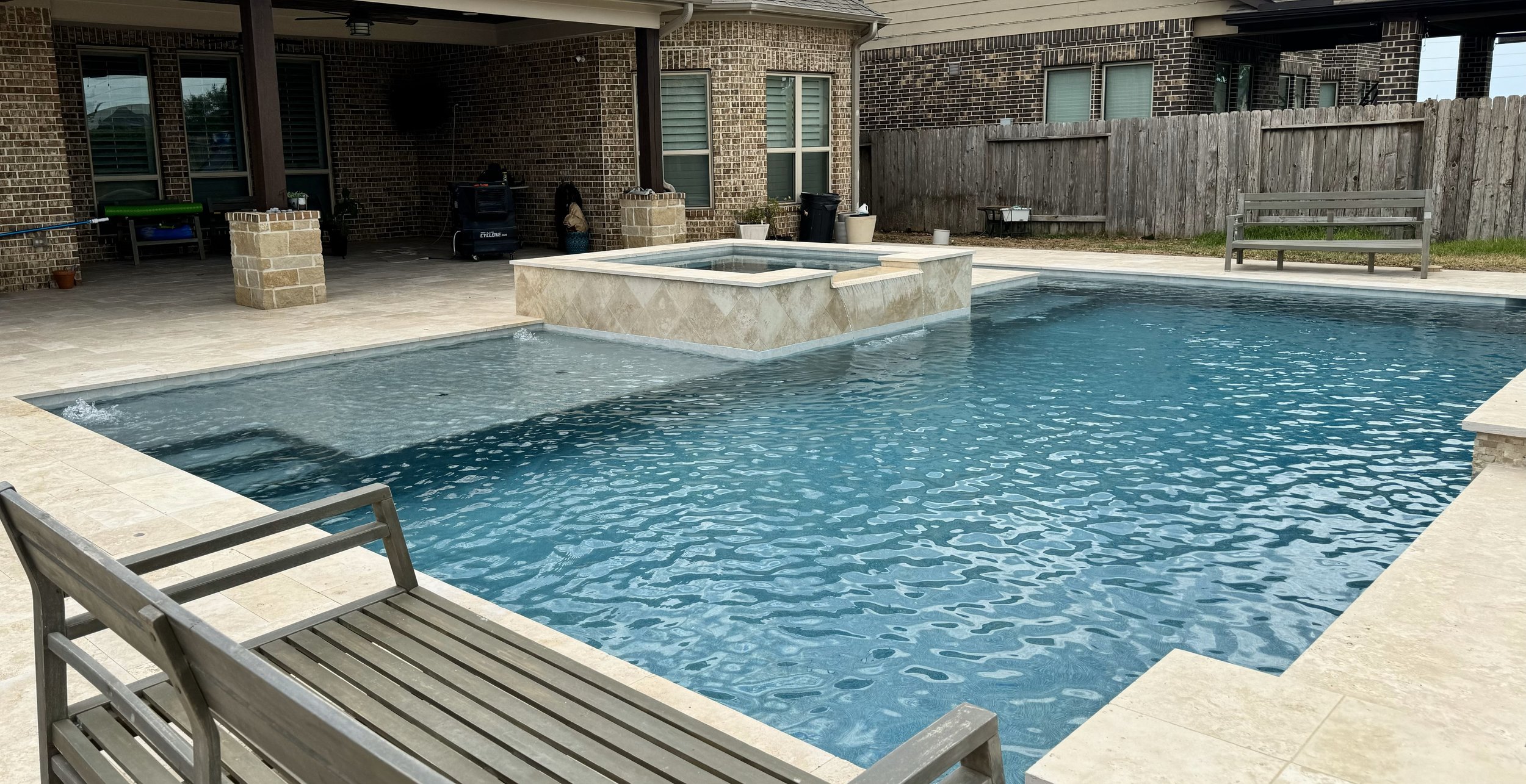 Backyard swimming pool with a raised hot tub integrated into the pool, surrounded by beige tile decking, with a wooden fence and brick house in the background. Benches are positioned around the area.