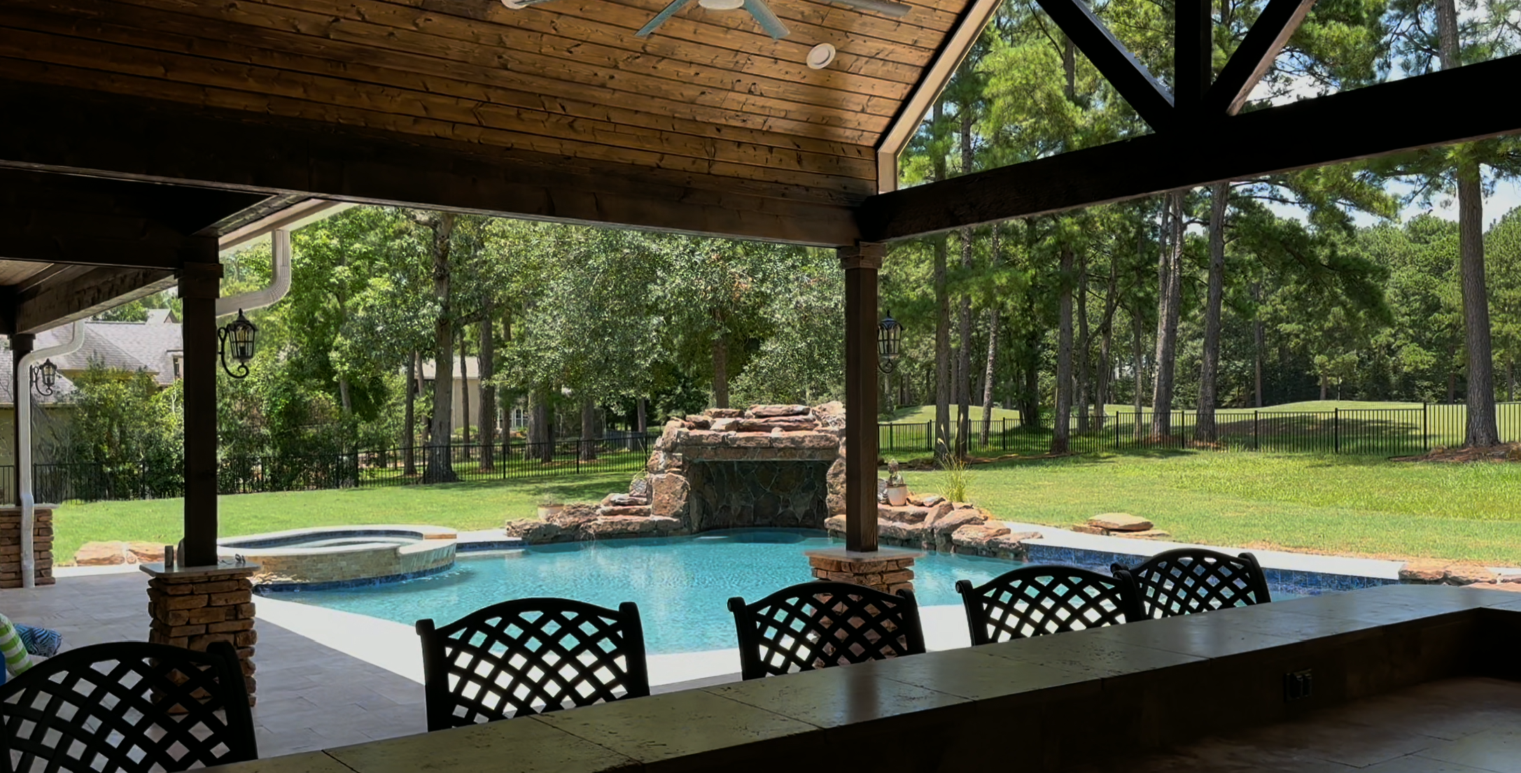 View from a covered patio overlooking a backyard with a swimming pool, rock waterfall feature, and green trees in the background.