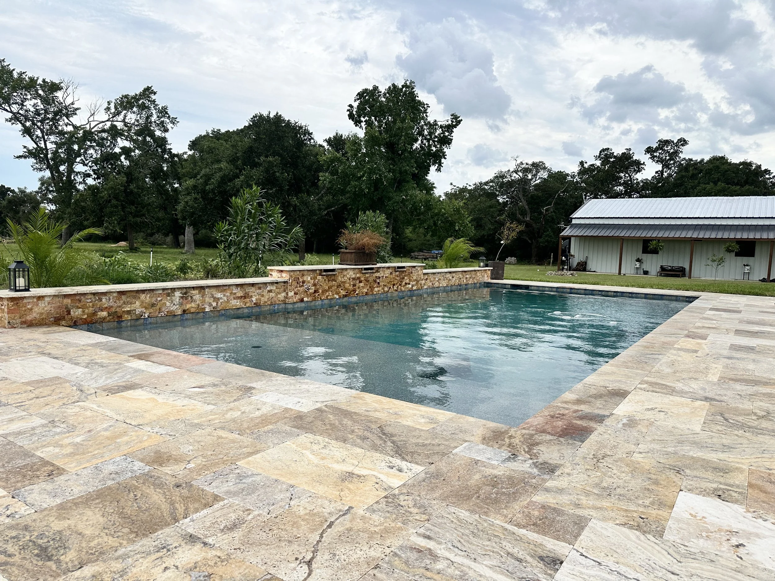 Outdoor backyard scene with a swimming pool, stone patio, lush green trees and plants, a small white building with a gray roof, and cloudy sky.