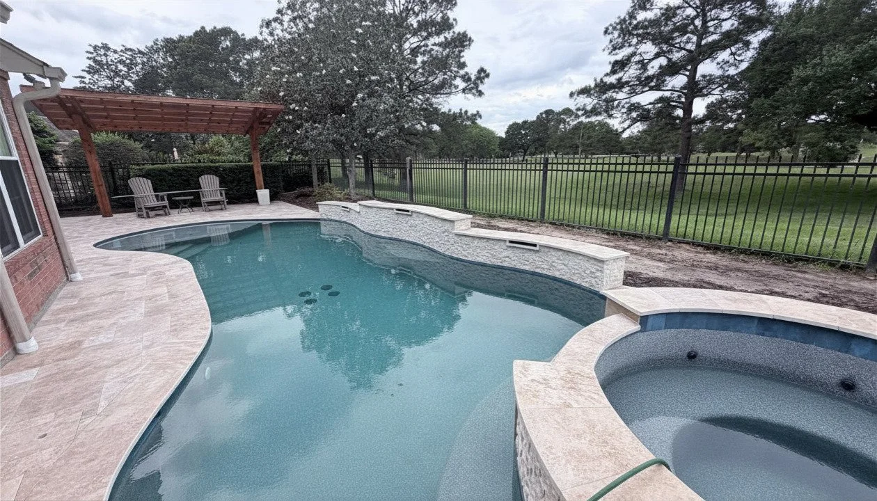 A backyard swimming pool with a hot tub, surrounded by a paved deck and a black metal fence, with trees and grass in the background under a cloudy sky.