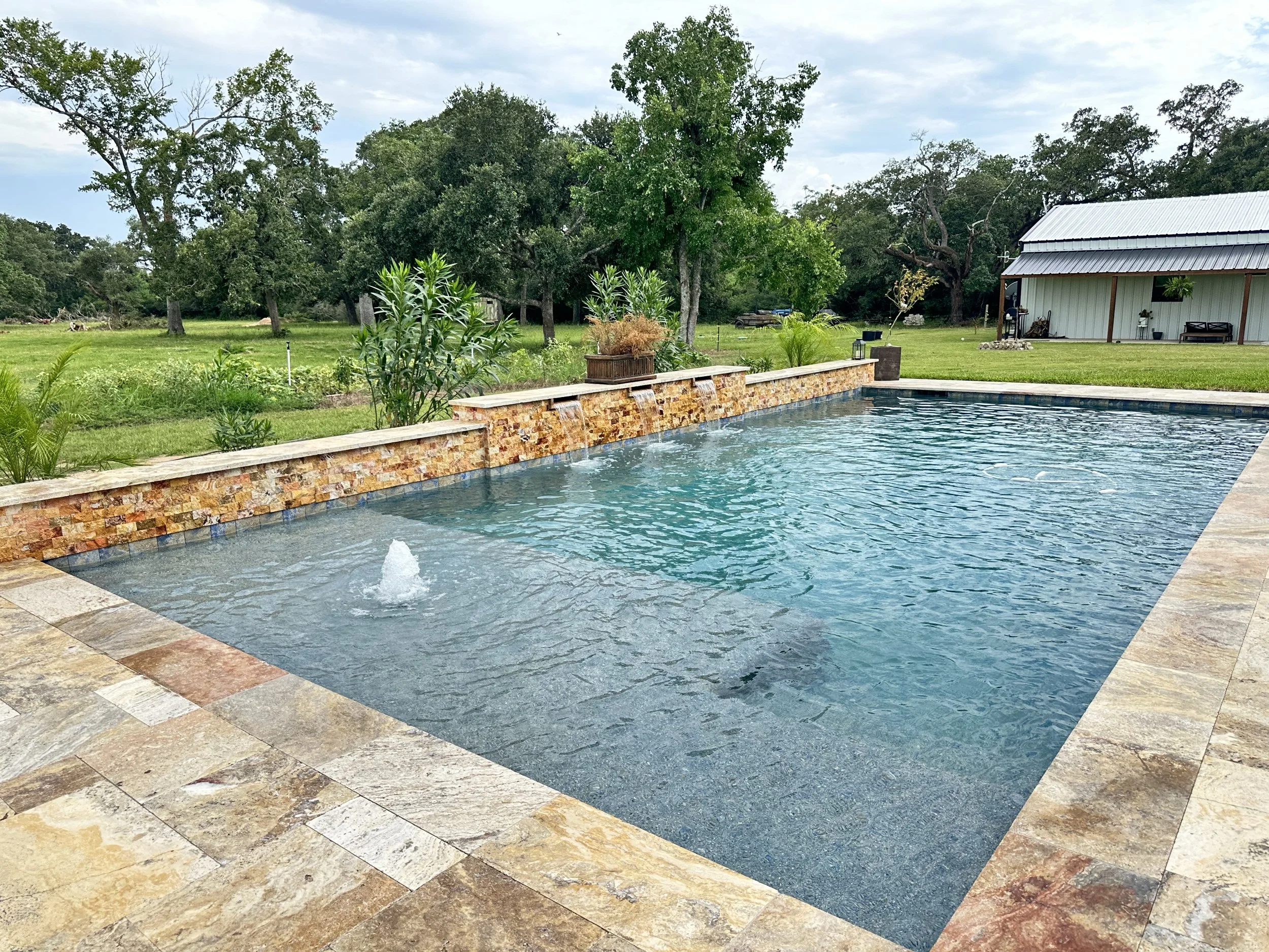 Outdoor scene with a swimming pool in the foreground, a lush grassy yard with trees, a barn, and cloudy sky in the background.