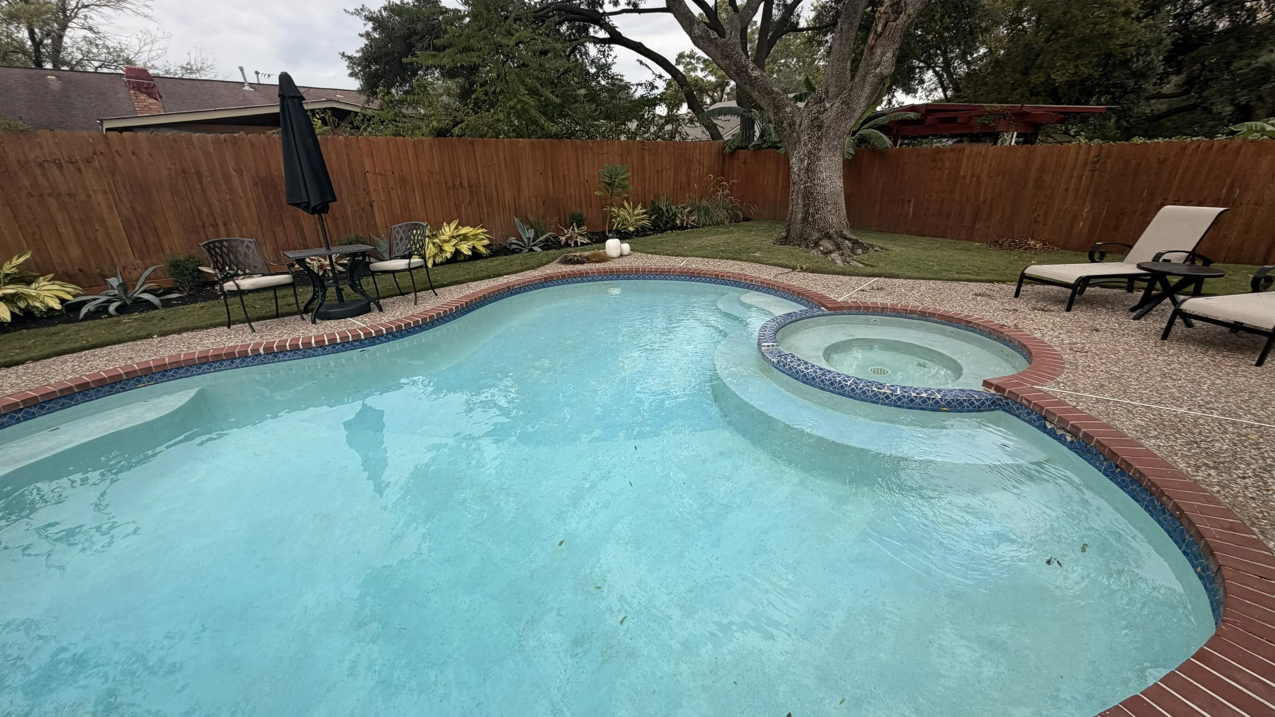 Backyard with swimming pool and hot tub, surrounded by a wooden fence, trees, garden plants, lounge chairs, table with chairs, and an umbrella.