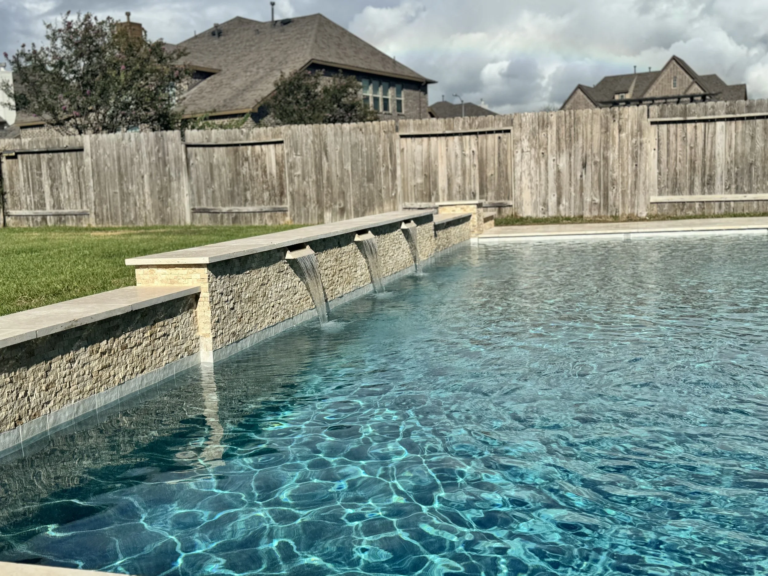 A swimming pool with clear blue water, stone edge, and a waterfall feature, surrounded by a grassy area and a wooden fence. Houses are visible in the background under a cloudy sky.