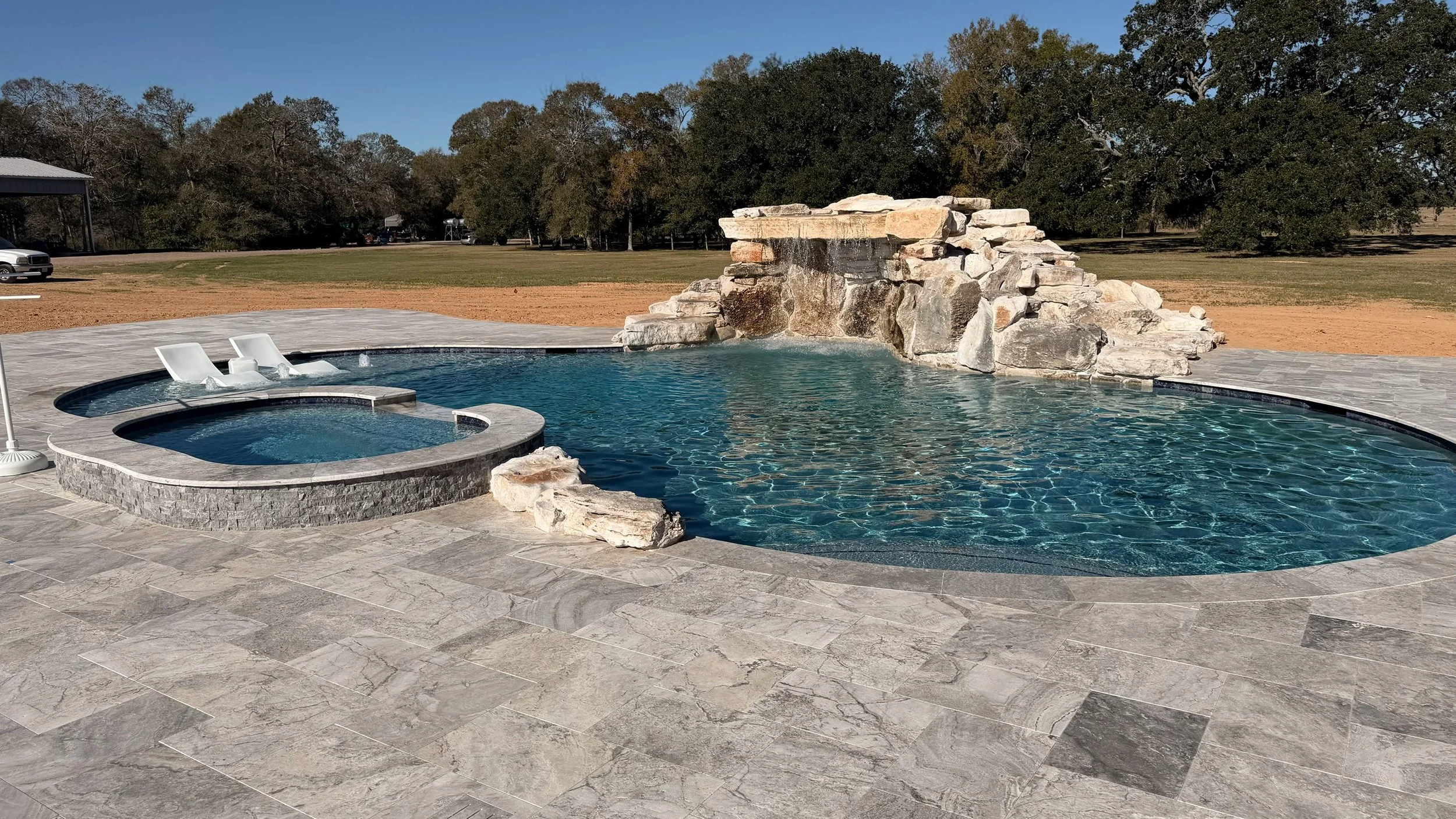 An outdoor swimming pool with a stone waterfall feature, adjacent hot tub, and surrounding paved area, with a grassy field and trees in the background under a clear blue sky.
