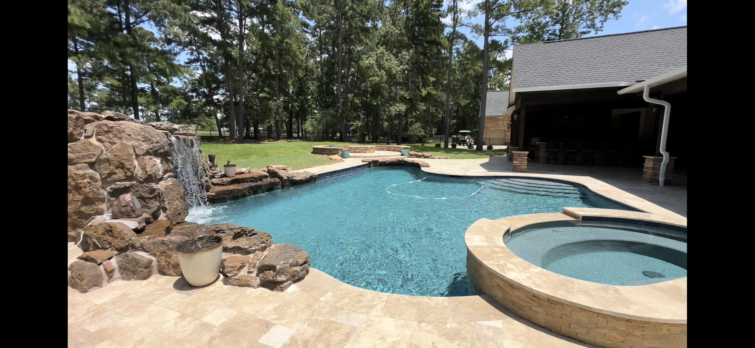 A backyard with a swimming pool and a hot tub, surrounded by a stone deck and a waterfall feature with rocks and water flowing into the pool. Trees and a cleared grassy area are visible in the background, and a covered patio area is on the right side