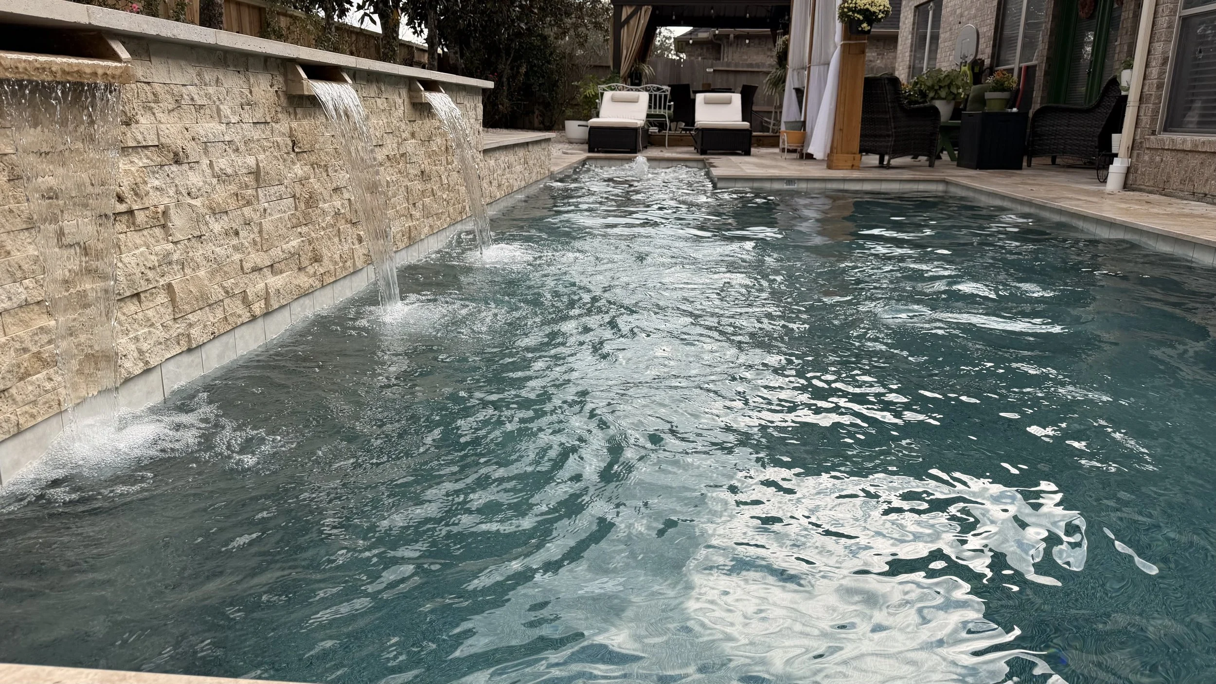 Backyard swimming pool with water features flowing from a stone wall, surrounded by outdoor furniture and potted plants.