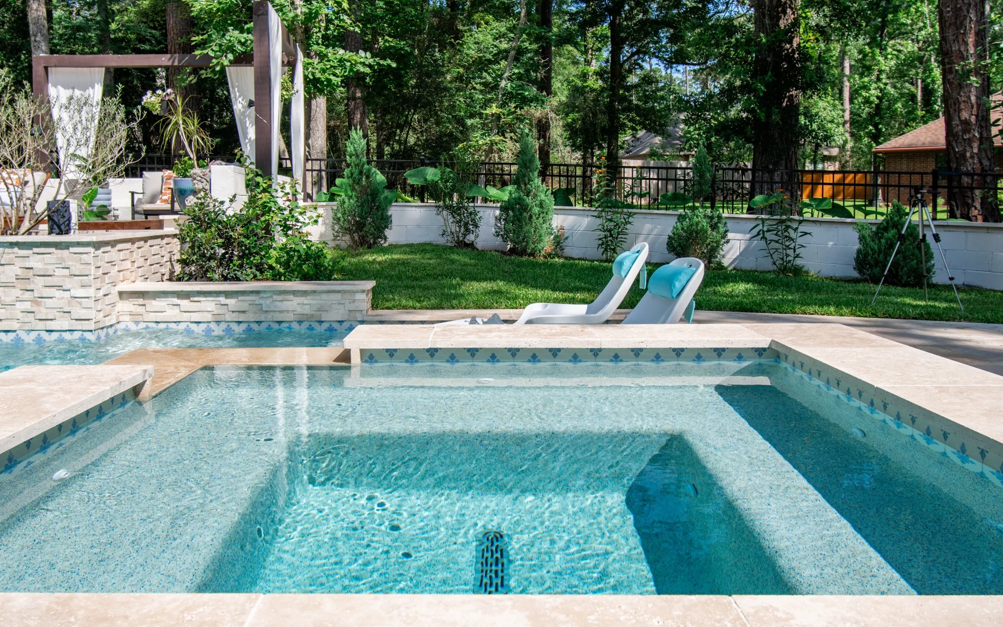 A backyard with a swimming pool, surrounded by greenery, trees, and a fence, with two lounge chairs on the pool deck.