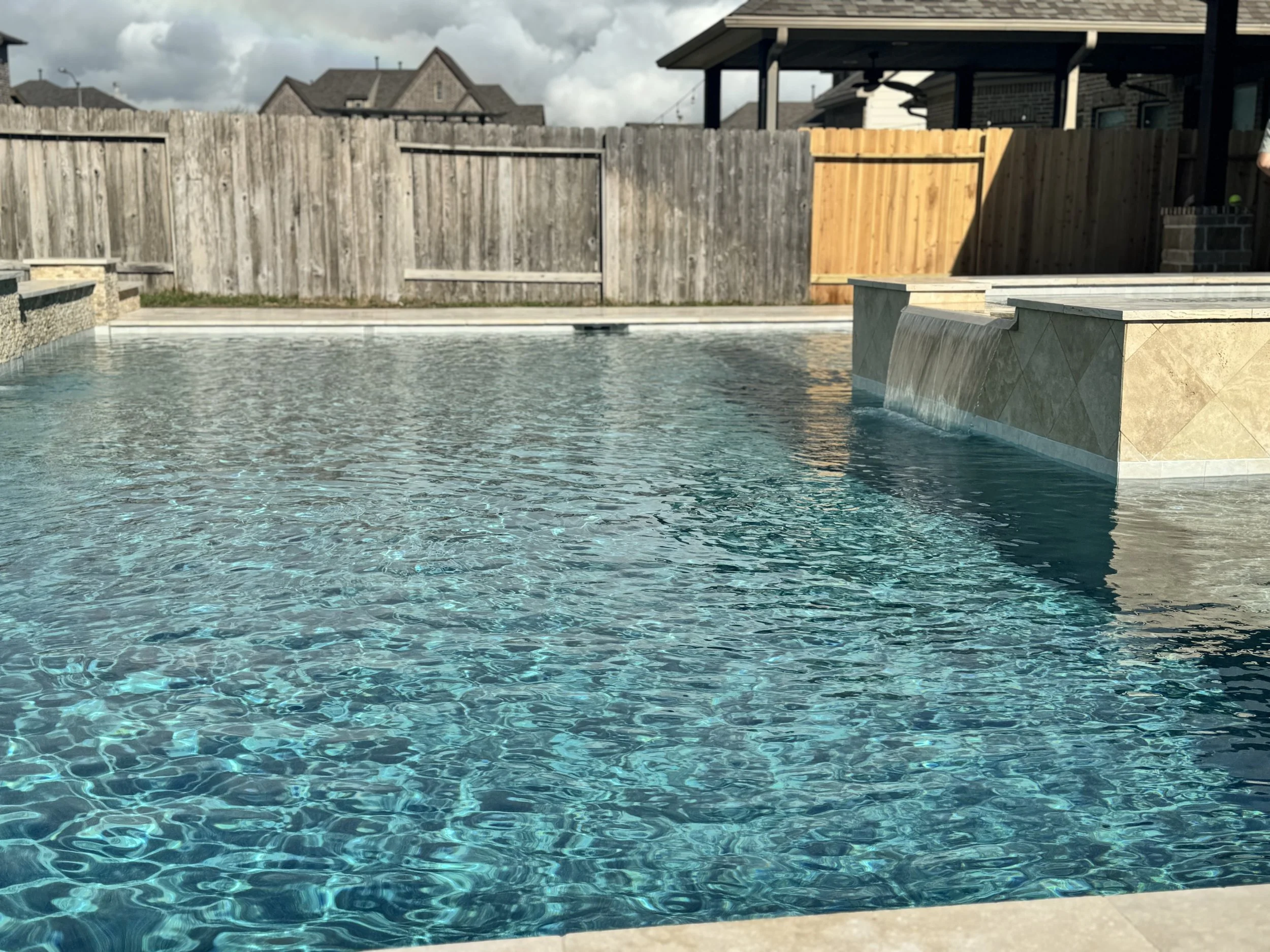 A backyard swimming pool with clear water, surrounded by a wooden fence. There is a section of the fence with natural wood color and another section painted dark. A house and cloudy sky are visible in the background.