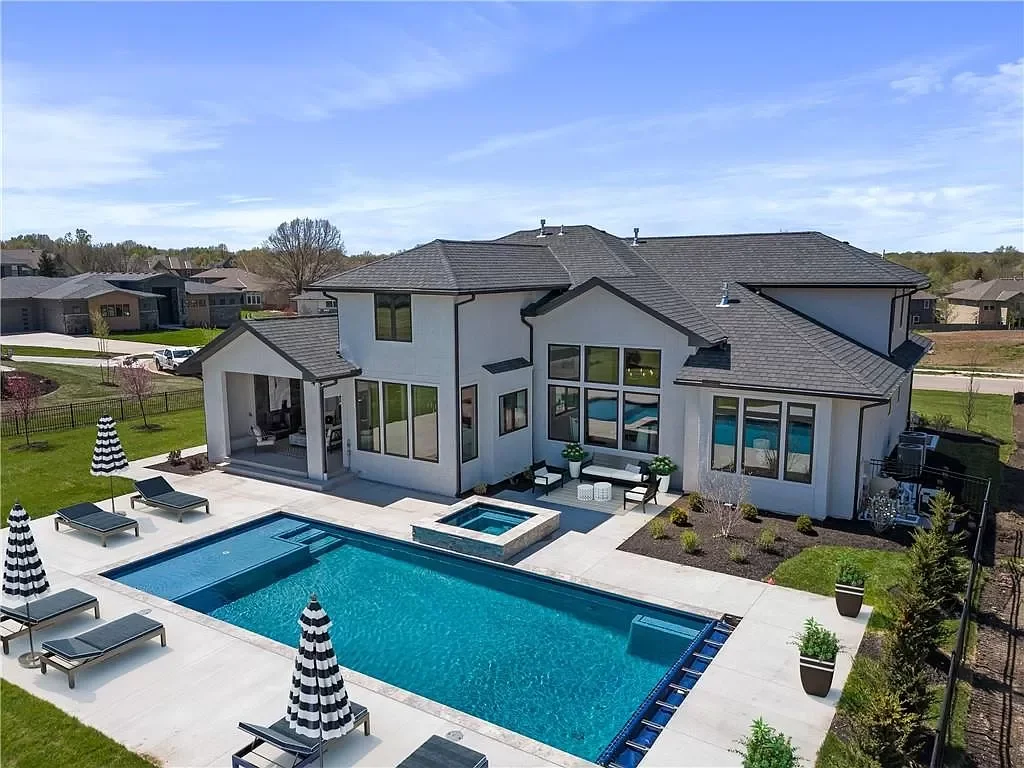 A modern two-story house with a backyard swimming pool, lounge chairs, umbrellas, and outdoor seating area under a clear blue sky.